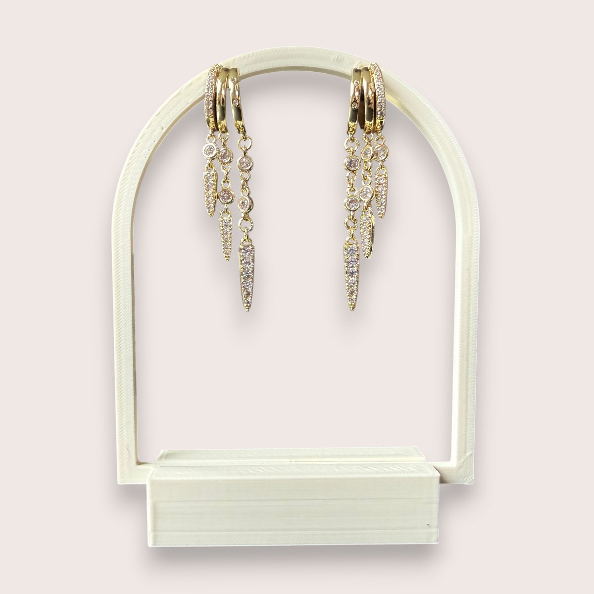 Nitelief gold hoop earrings with dangling crystal-encrusted spike charms on a white arched display stand.