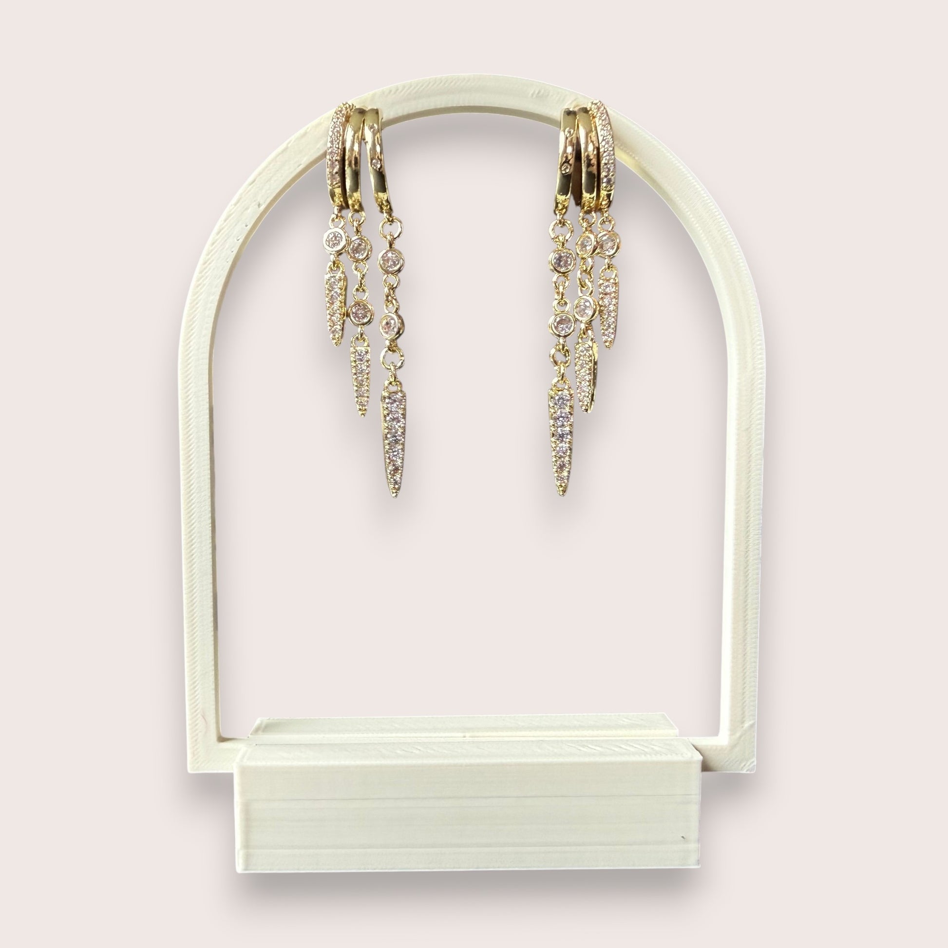 Nitelief gold hoop earrings with dangling crystal-encrusted spike charms on a white arched display stand.